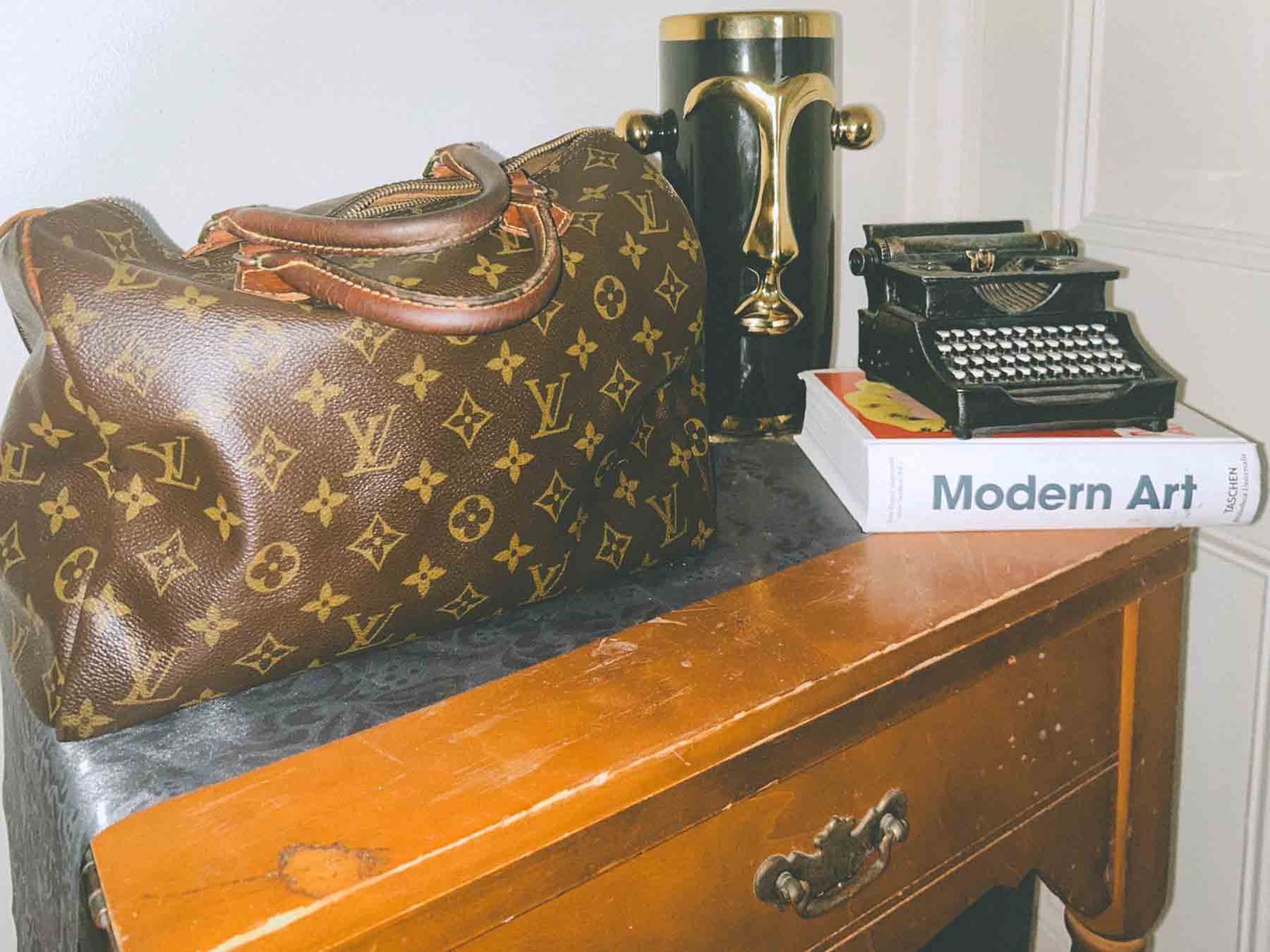 Inside My Closet — styled Louis Vuitton Speedy bag resting on a vintage wood table beside a Modern Art book, black vase, and mini typewriter, reflecting bohemian luxury and artistic living.