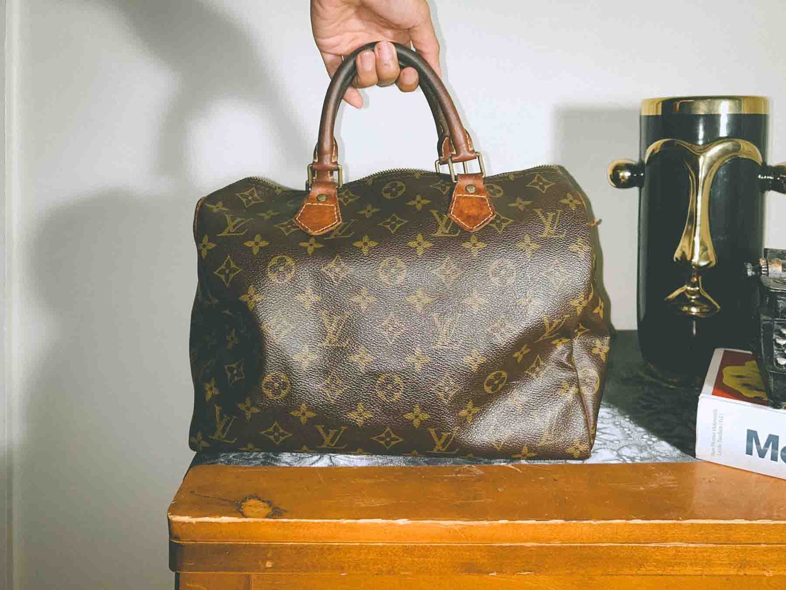 Inside My Closet — vintage Louis Vuitton Speedy bag being held by its handle on a wooden table beside a sculptural gold vase, symbolizing timeless style and self-expression.