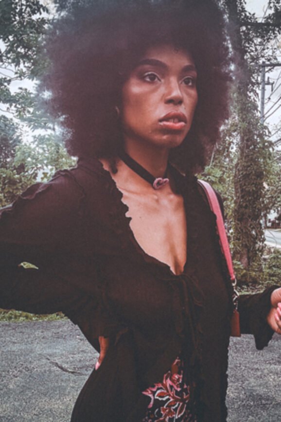 Woman standing in a parking lot wearing a gothic-inspired black ruffle blouse layered over a magenta floral dress, styled with a velvet choker and natural afro.