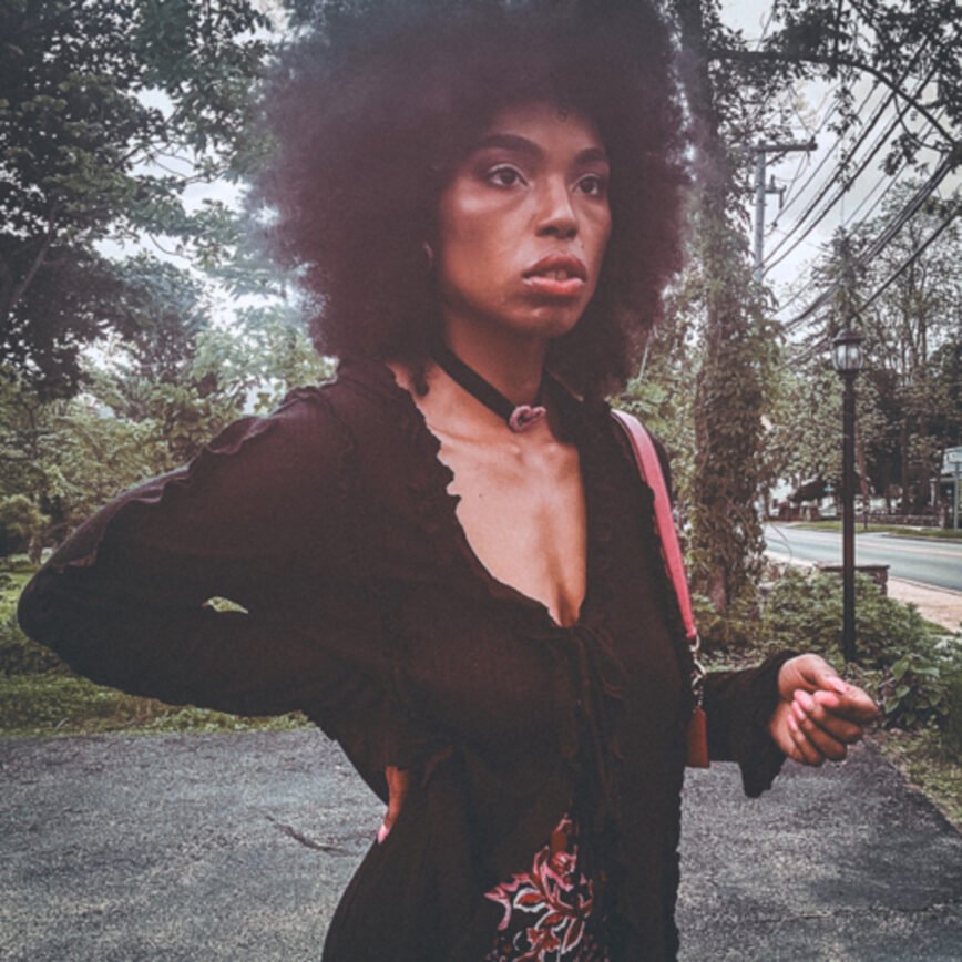 Woman standing in a parking lot wearing a gothic-inspired black ruffle blouse layered over a magenta floral dress, styled with a velvet choker and natural afro.