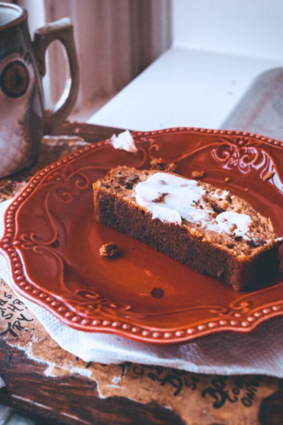 A cozy breakfast scene featuring a slice of buttered sweet potato bread on a red ornate plate, placed on a napkin over a rustic wooden board. A ceramic owl-shaped mug sits nearby, with a butter knife resting on the board. Soft natural light filters through an old window, creating a warm, inviting atmosphere.