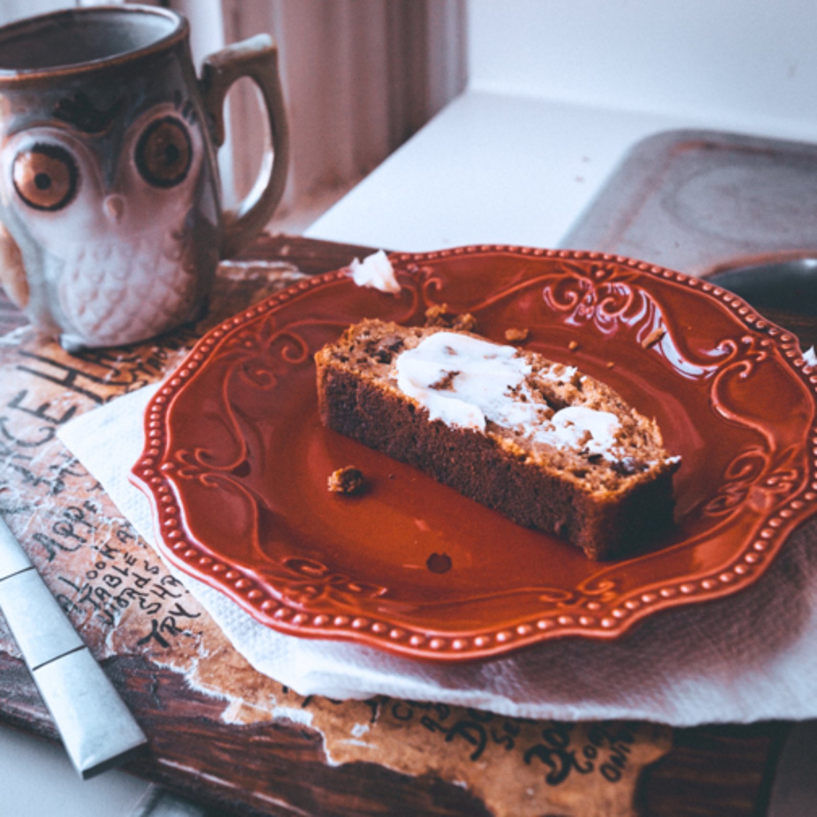 A cozy breakfast scene featuring a slice of buttered sweet potato bread on a red ornate plate, placed on a napkin over a rustic wooden board. A ceramic owl-shaped mug sits nearby, with a butter knife resting on the board. Soft natural light filters through an old window, creating a warm, inviting atmosphere.