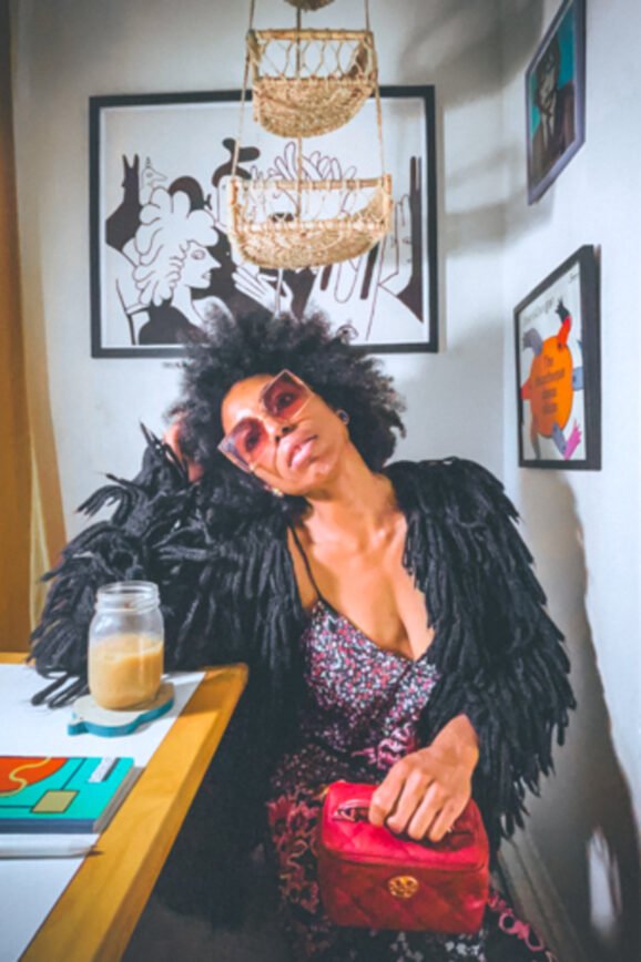 Woman with a natural afro hairstyle and oversized pink sunglasses sits at a white kitchenette table with an iced coffee, resting her head on her hand. She wears a black fringe jacket over a patterned dress and holds a red handbag. The room is decorated with modern black-and-white art prints, a woven hanging basket, and warm lighting from golden curtains, creating a cozy, bohemian vibe.