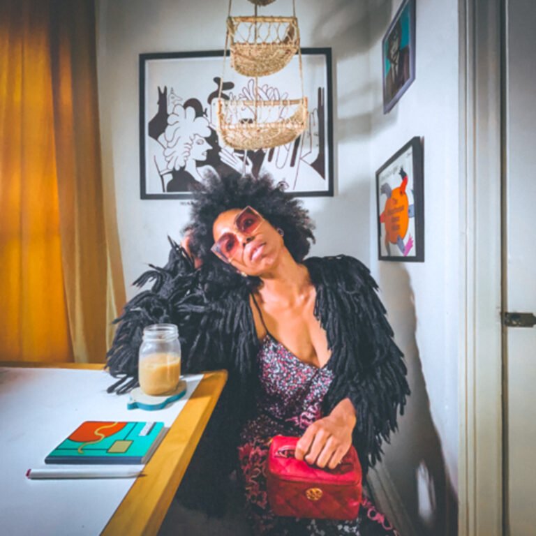Woman with a natural afro hairstyle and oversized pink sunglasses sits at a white kitchenette table with an iced coffee, resting her head on her hand. She wears a black fringe jacket over a patterned dress and holds a red handbag. The room is decorated with modern black-and-white art prints, a woven hanging basket, and warm lighting from golden curtains, creating a cozy, bohemian vibe.