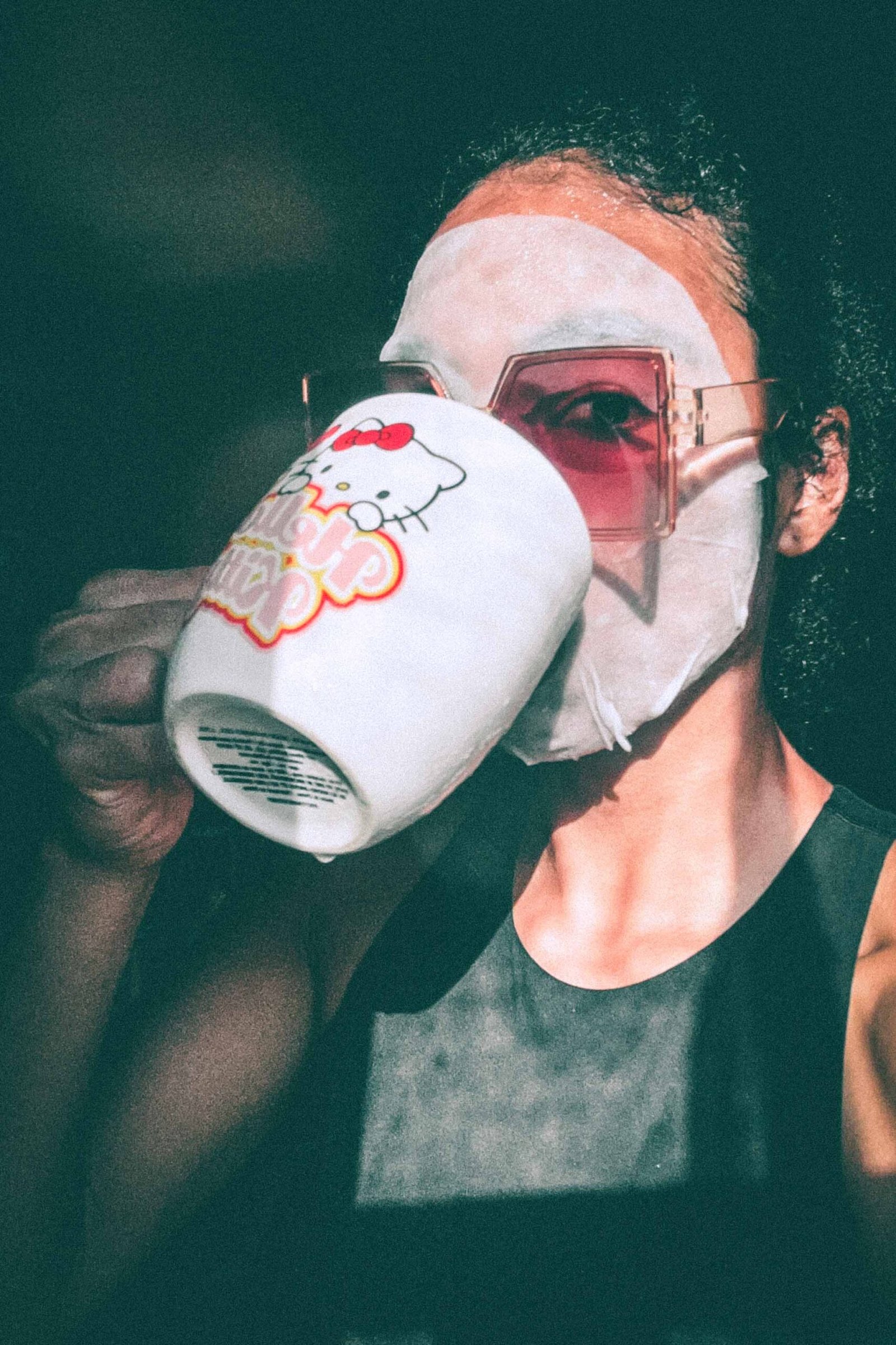 Woman wearing a sheet mask and oversized sunglasses while sipping from a Hello Kitty mug during her skincare routine.