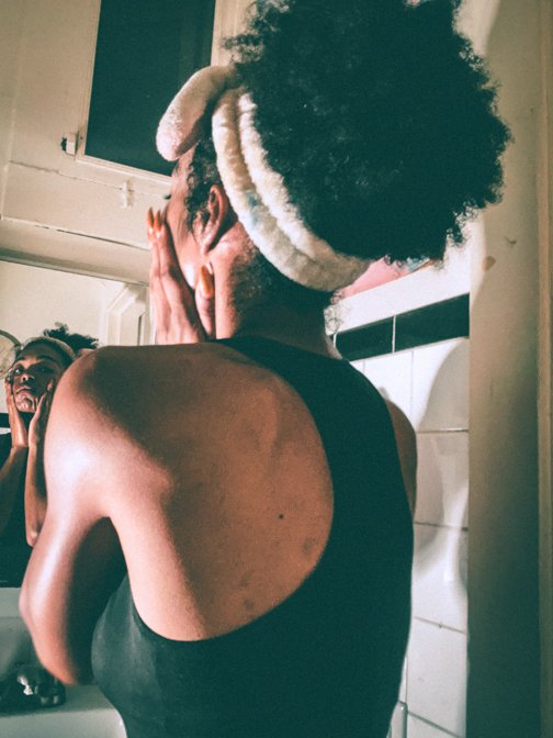 Back view of a woman in a tank top applying skincare in a softly lit bathroom with a focus on her self-care ritual.
