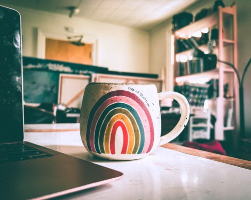 Rainbow mug labeled 'Cup of Gratitude' on white table beside laptop in cozy bohemian home with bookshelves in background.