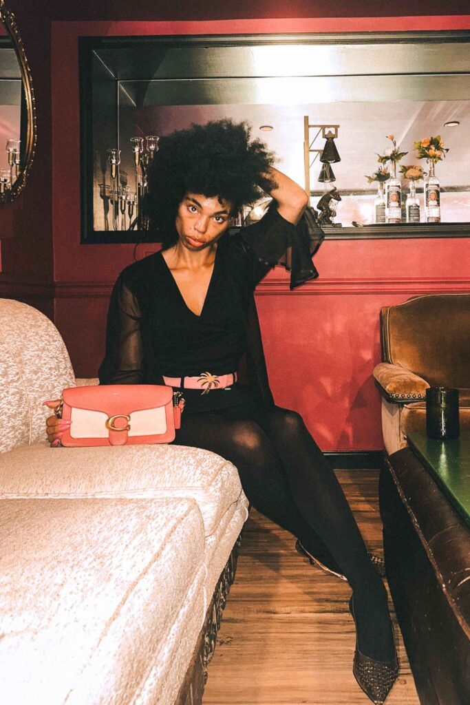 Woman in black dress with sheer sleeves, pink Palm Angels belt, and coral Coach clutch sitting on cream sofa in pink-walled lounge.