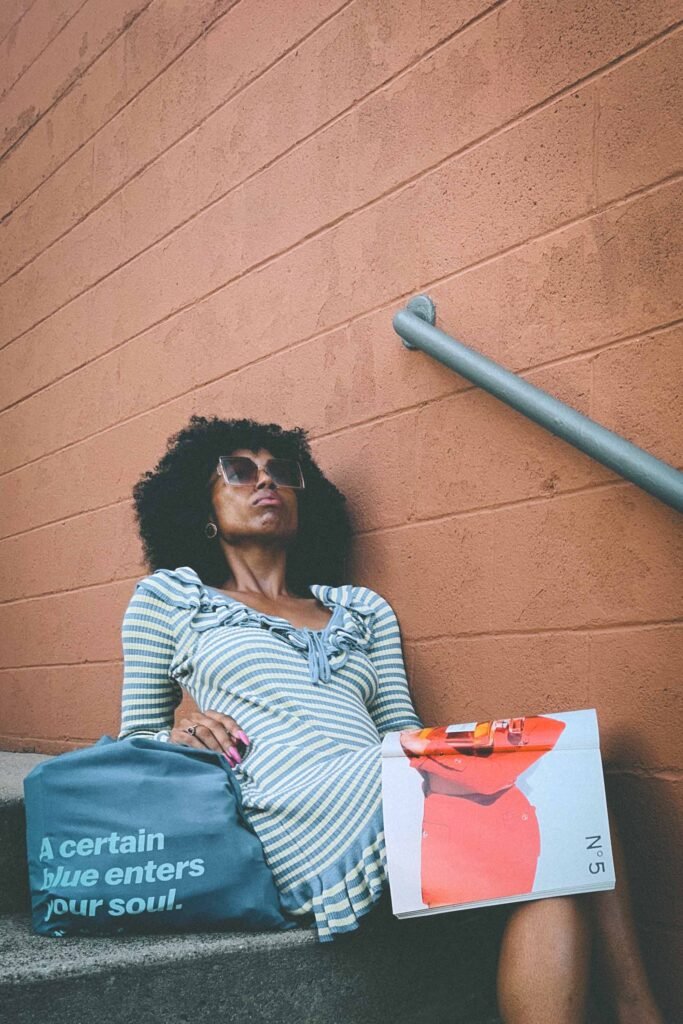 Fashionable woman in blue and yellow striped ruffle dress sitting on outdoor stairs with blue tote bag and open fashion magazine.