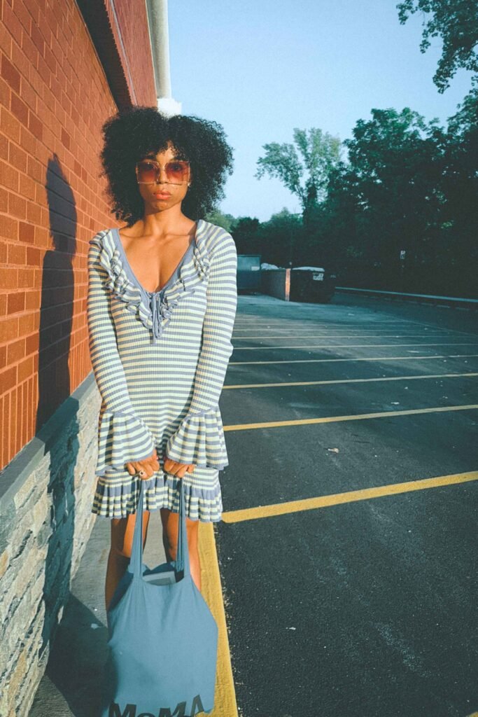 Woman standing in blue and yellow striped ruffle dress with flared sleeves holding blue MoMA tote bag.