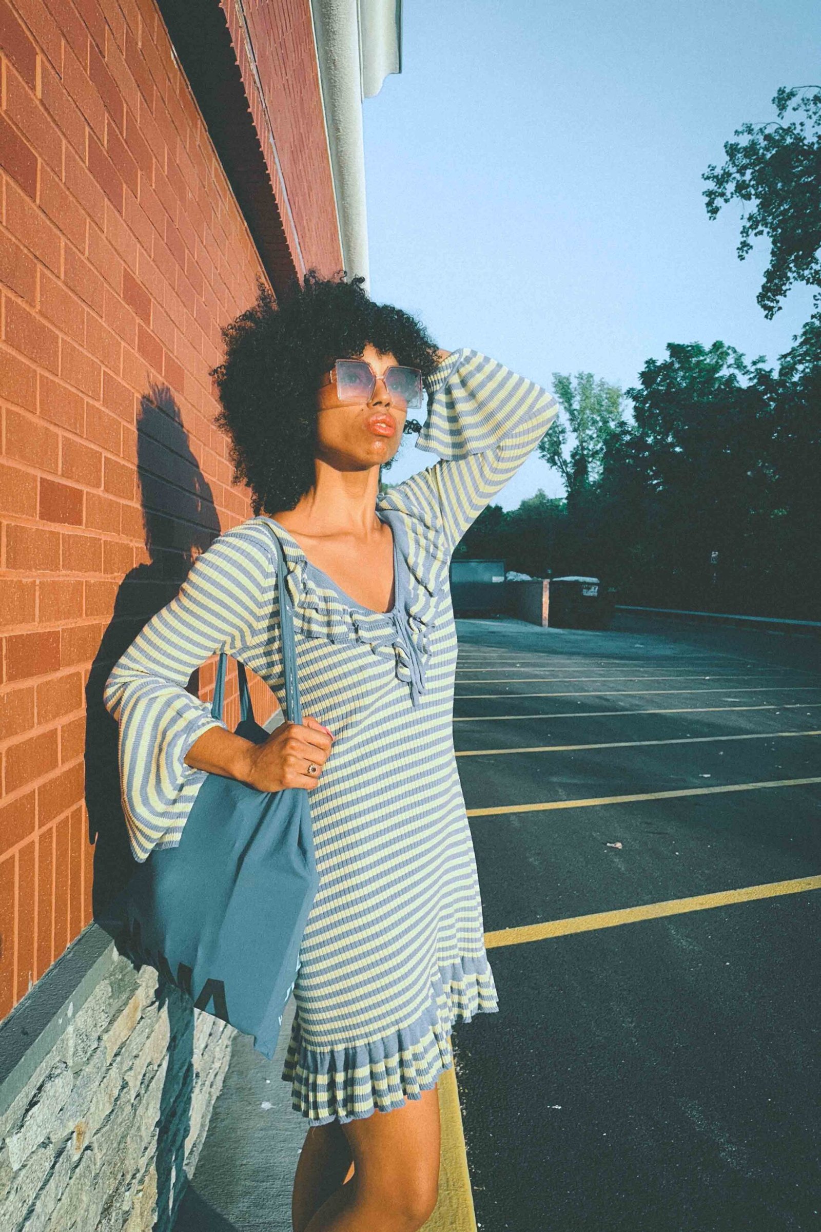 Woman in blue and yellow striped ruffle dress with flared sleeves holding blue tote bag in golden hour light against brick wall.