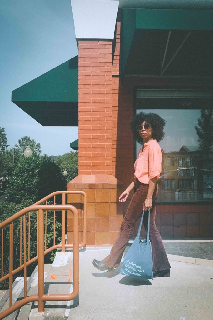 Woman in coral blouse and brown corduroy pants walking past brick building carrying blue Henri Matisse tote bag and wearing black clogs.