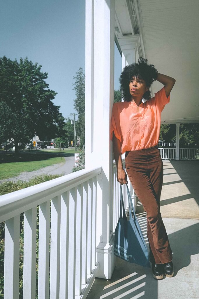 Woman in loose coral blouse and high-waisted brown corduroy pants holding blue tote bag on sunlit porch.