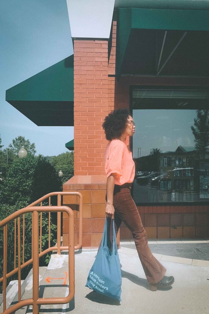 Stylish woman wearing coral blouse, brown corduroy pants, and black clogs carrying blue Henri Matisse tote bag past brick building.