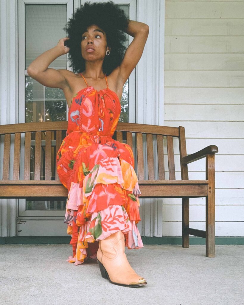Woman wearing orange Farm Rio maxi dress with ruffle tiers and tan cowboy boots, styled on a porch for bohemian fashion.