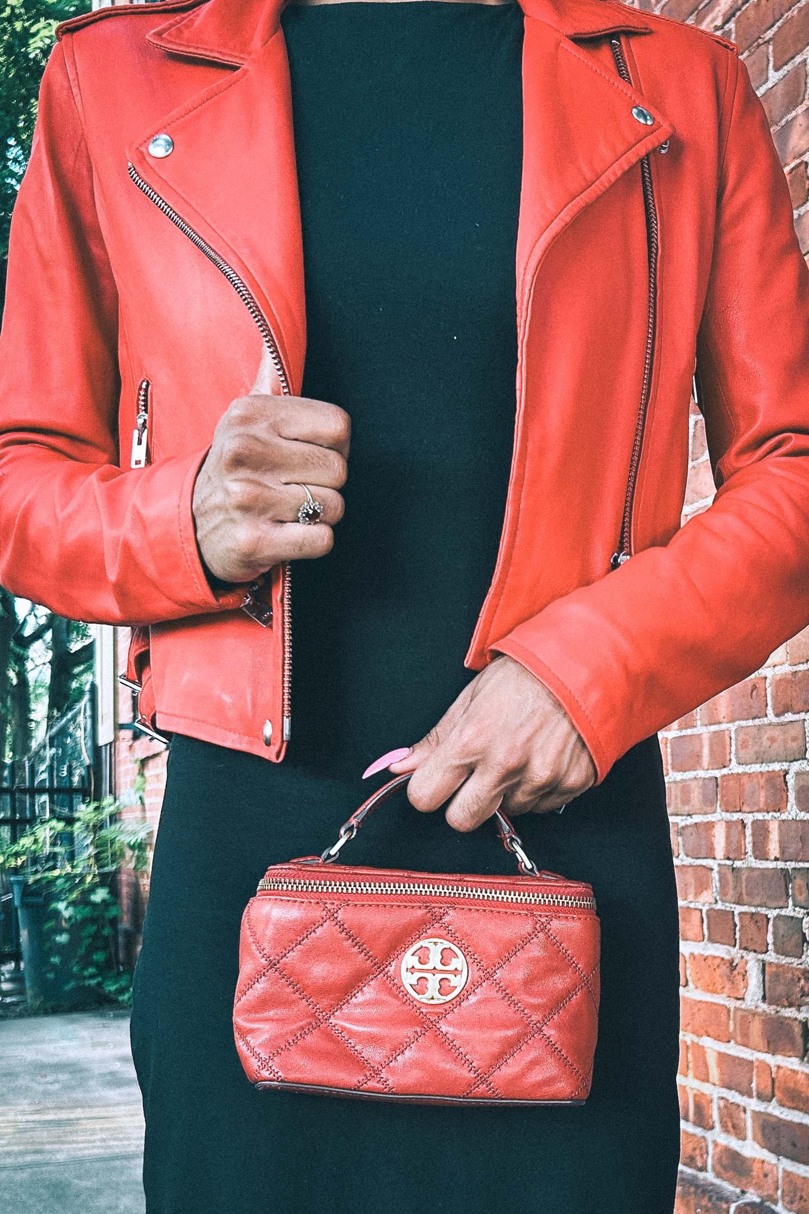Close-up of woman wearing a red leather moto jacket over a black dress, holding a small quilted Tory Burch handbag, urban brick wall background.