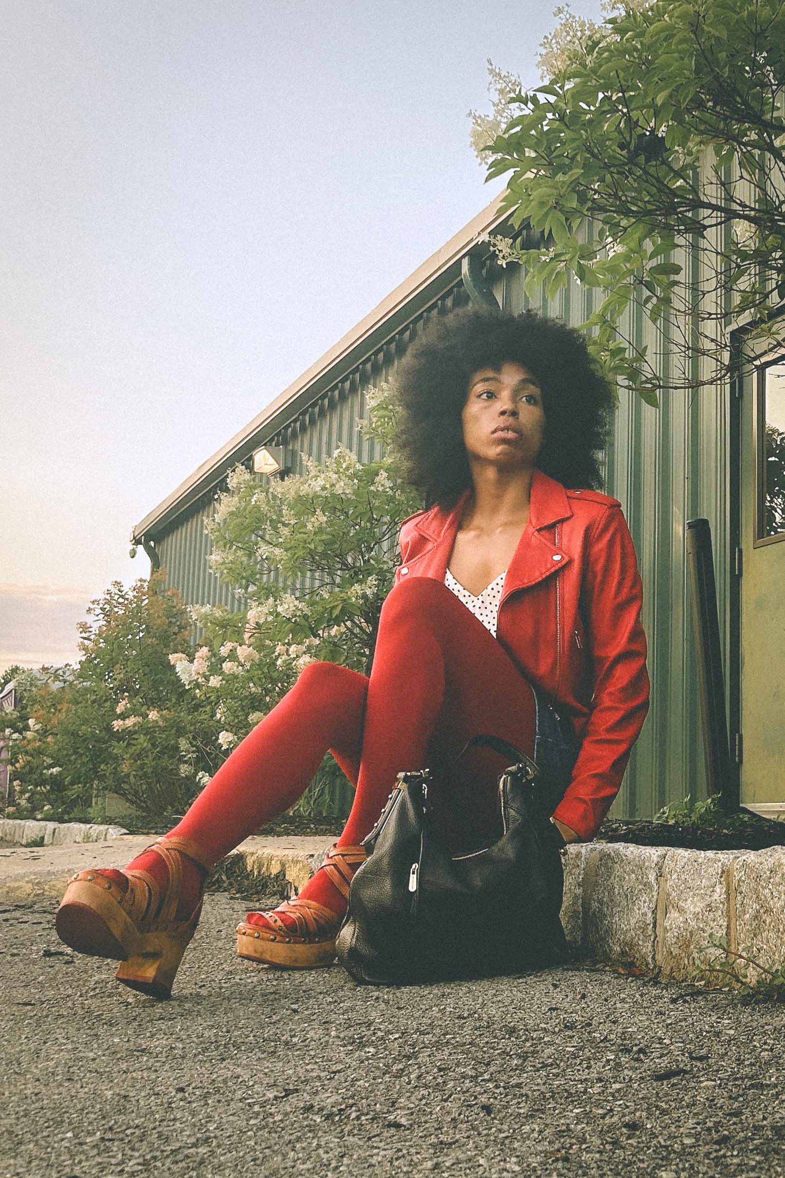 Afro-haired woman in a bold red leather jacket, red tights, and platform sandals sitting on a curb with a black leather bag, shot at golden hour against a green building.