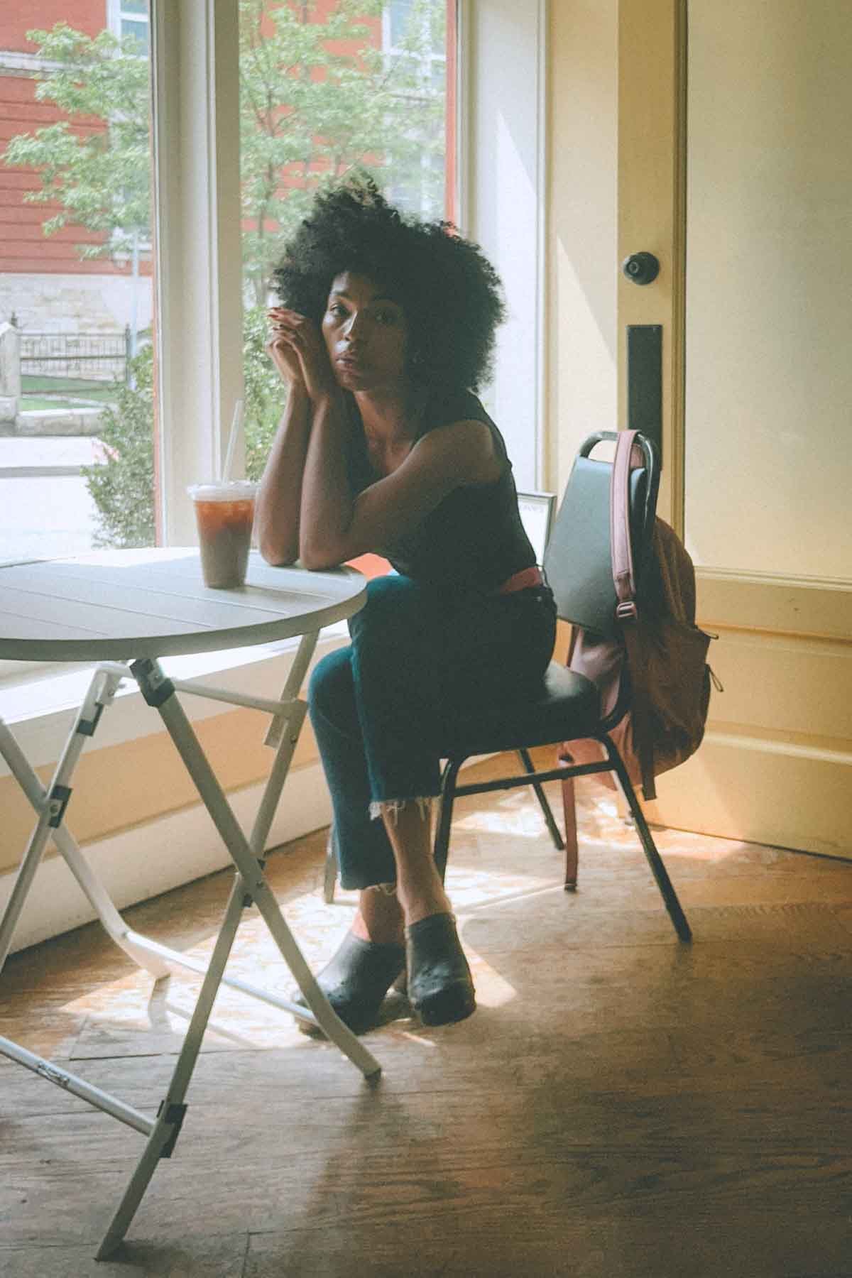 Woman sitting thoughtfully by a café window with iced coffee, sunlight spilling across the floor — a quiet moment of soft productivity and creative reflection.