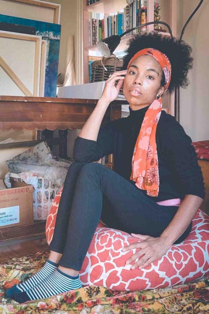 Creative portrait of a woman seated on a floor cushion in her art-filled apartment, surrounded by books and canvases — embodying artistic renewal.