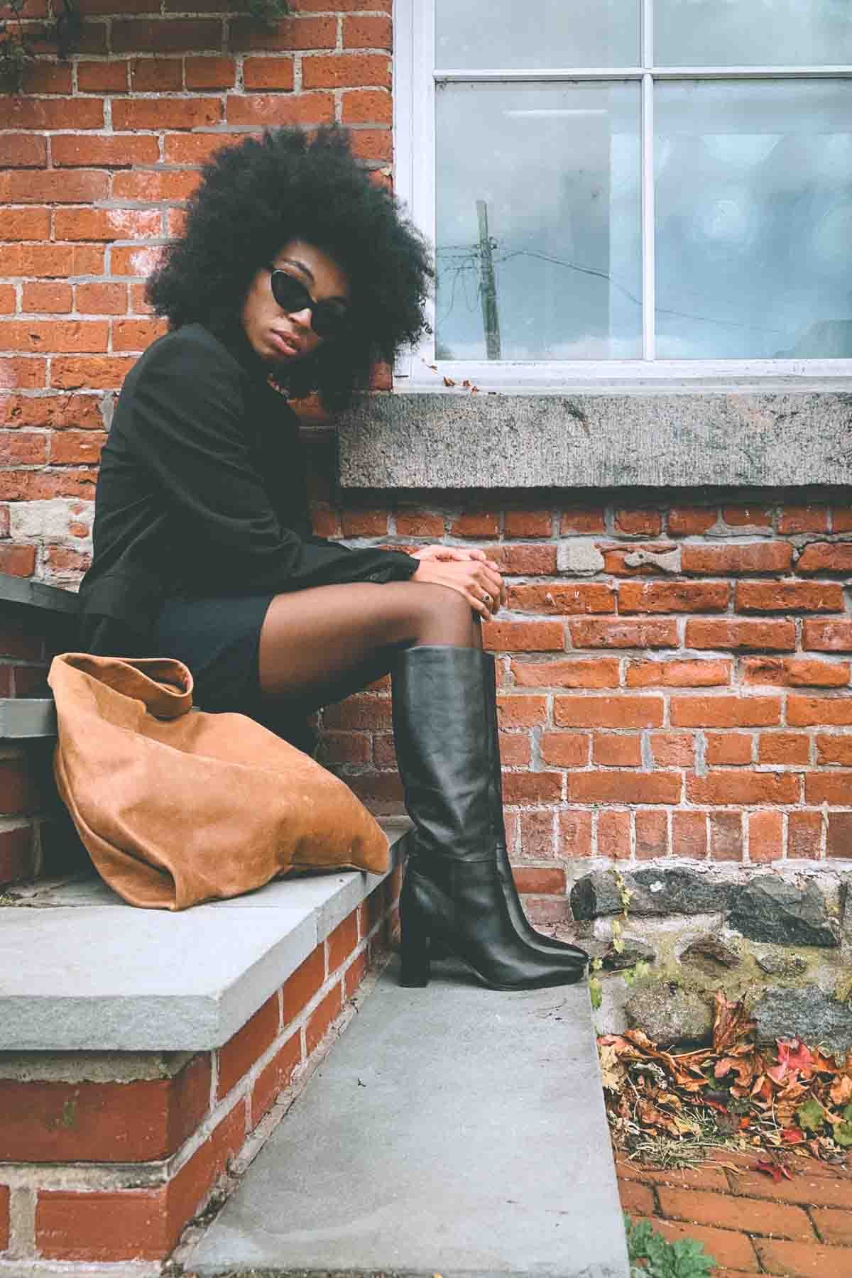 Bohemian fashion blogger in black blazer, mini skirt, and knee-high leather boots sitting on brick steps for fall outfit diary.