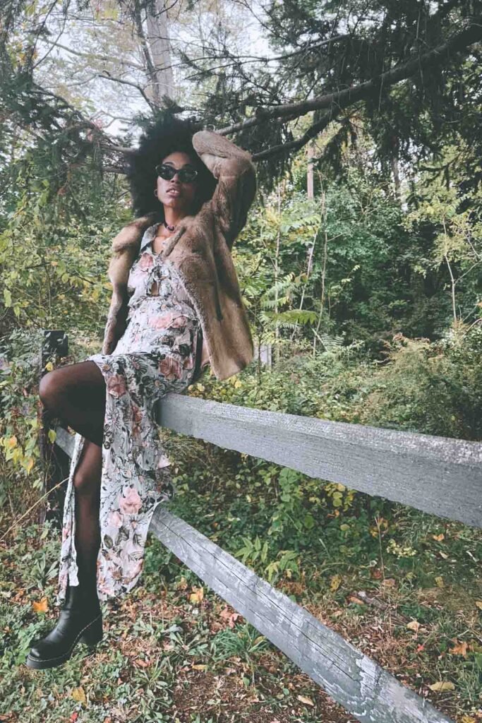 Bohemian fashion blogger sitting on a wooden fence in the woods wearing a vintage fur coat, floral maxi dress, and black platform boots for a fall “Inside My Closet” outfit shoot.