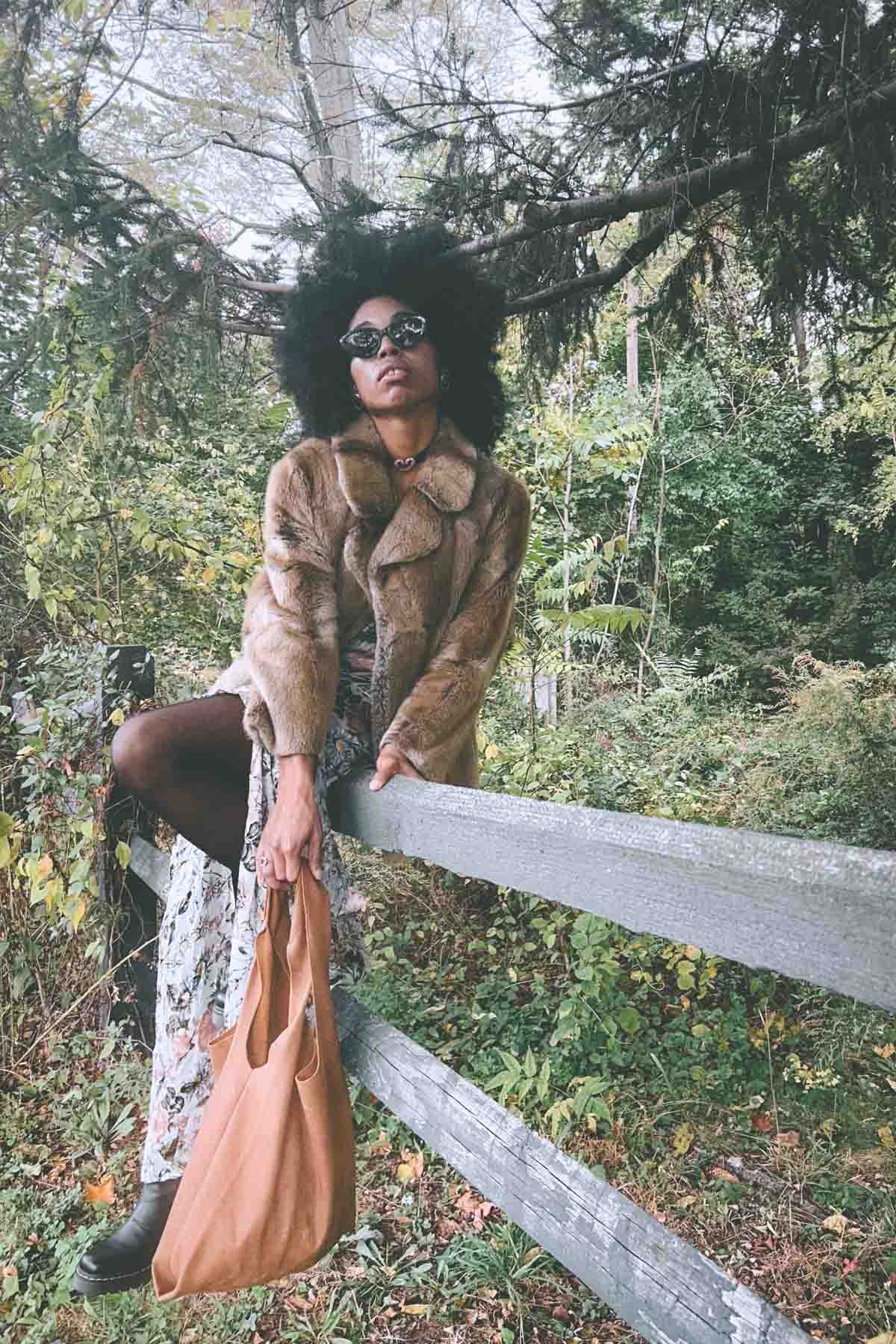 Bohemian blogger posing on a weathered fence in a forest, styled in a vintage fur coat, floral ruffled dress, black boots, and a suede tote bag for an autumn wardrobe editorial.