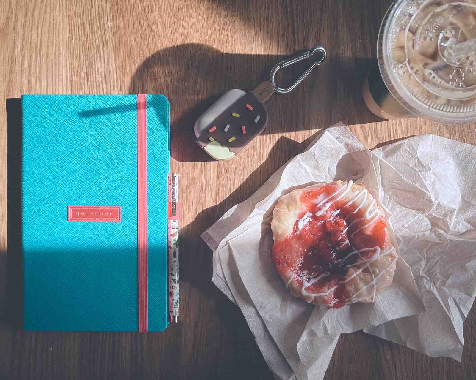 Flatlay of a turquoise journal, pen, iced coffee, and cherry danish pastry on a wooden table — cozy morning routine inspiration for writers and creatives.