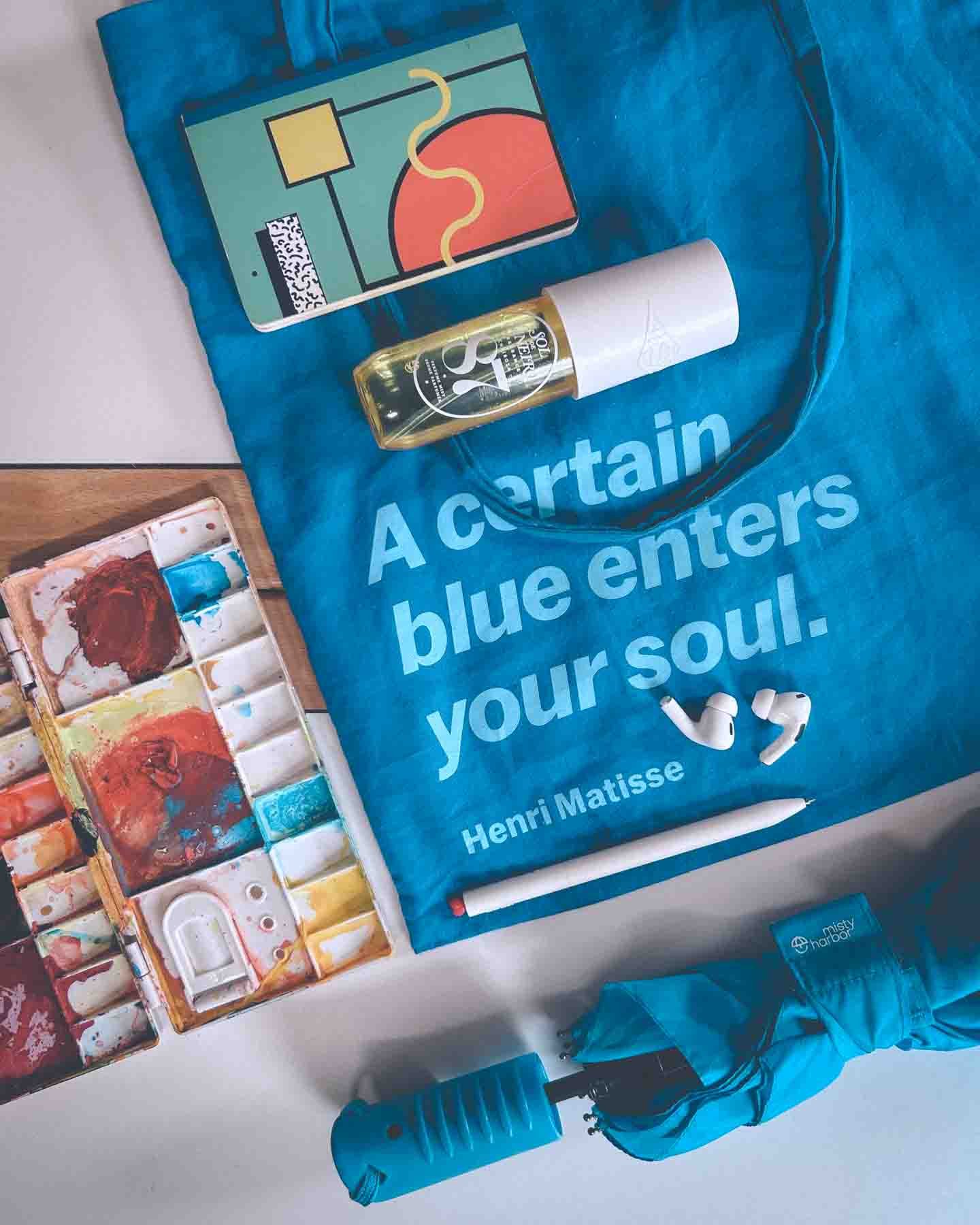 Flatlay of art supplies, Matisse tote bag, perfume, umbrella, and AirPods — bohemian essentials for a creative day on the go.