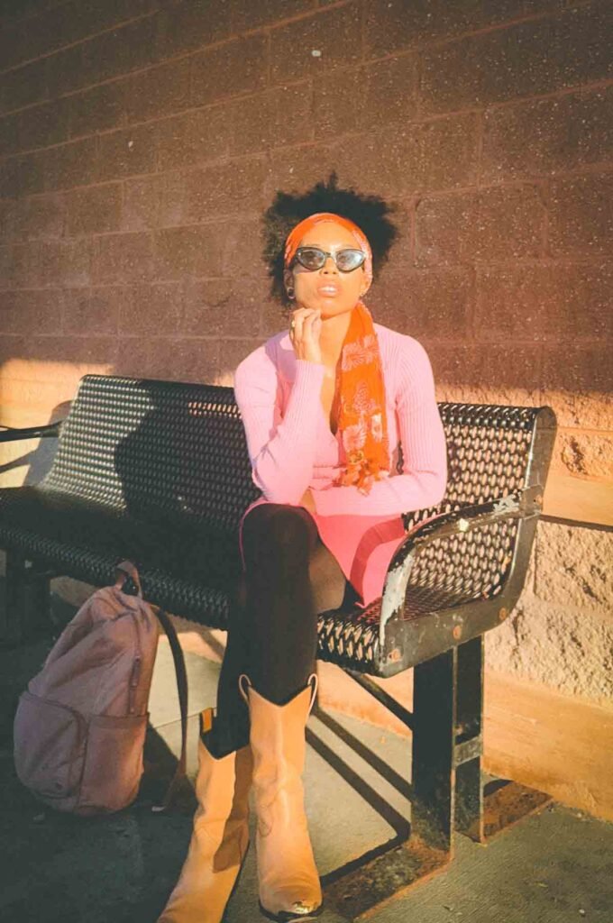 Woman sitting on a metal bench during golden hour wearing a pink ribbed top, mini skirt, orange scarf, and tan cowboy boots — bohemian fall fashion with vintage editorial sunlight.