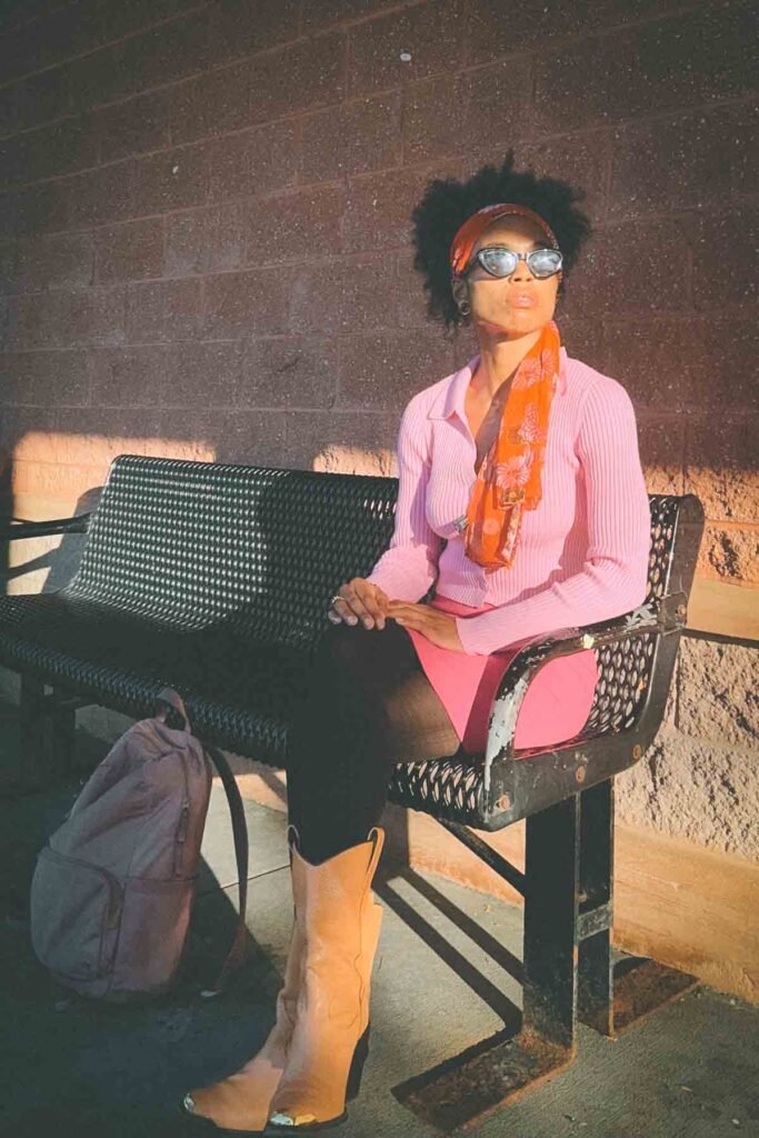 Woman in a pink ribbed top, mini skirt, and orange scarf sitting on a bench at golden hour — vintage-inspired bohemian fall outfit with cowboy boots and soft sunlight.