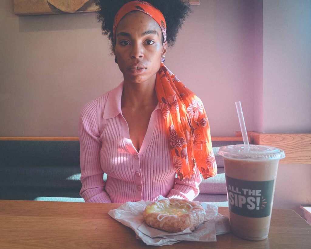 Bohemian lifestyle moment in a café—woman in a pink ribbed top with an orange floral scarf, sitting with an iced coffee and lemon pastry, capturing the essence of slow mornings, mindful rituals, and soft productivity.