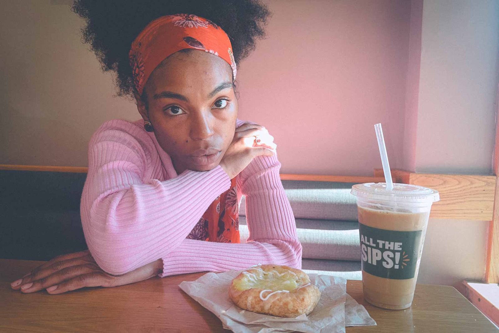 Portrait of a woman in a pink top and orange scarf gazing thoughtfully at the camera with coffee and pastry — balancing beauty and reflection in everyday life.