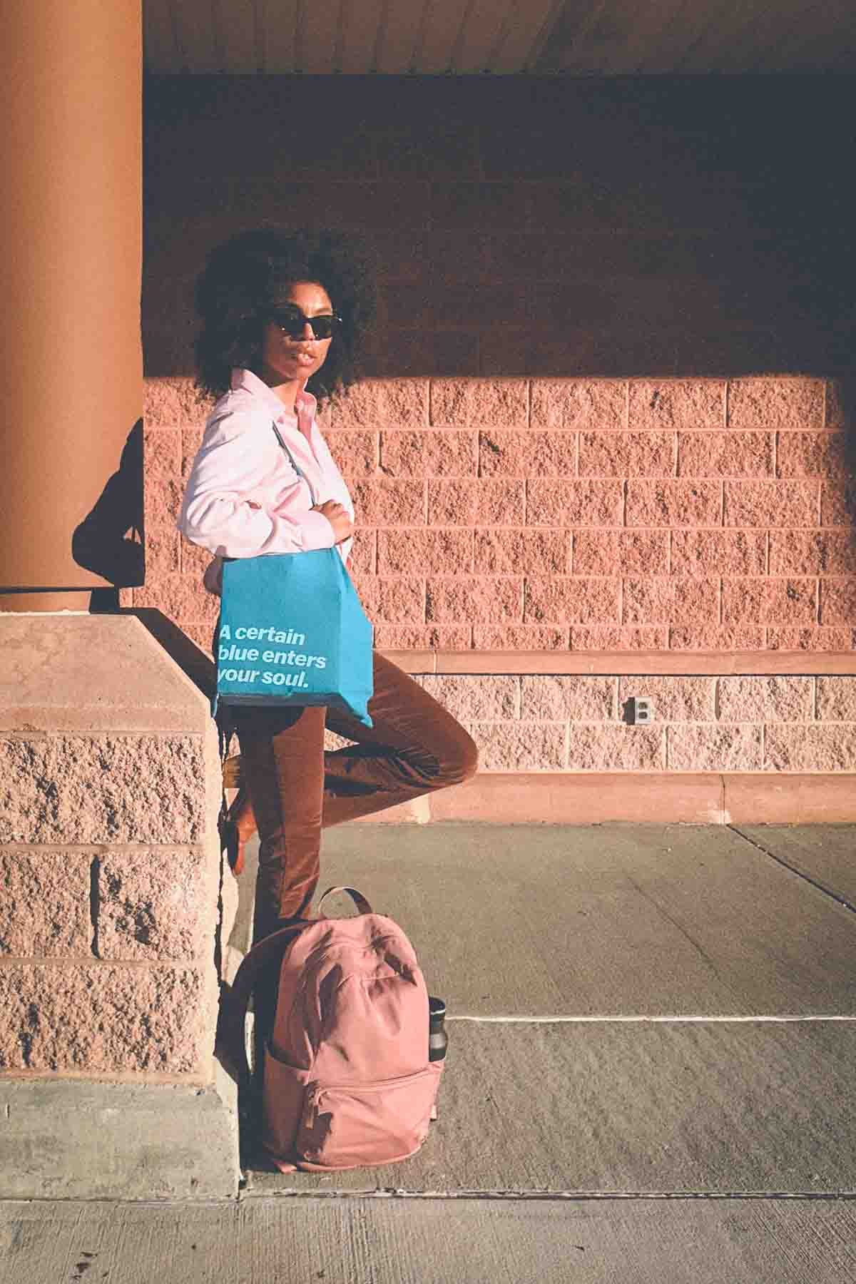 Woman standing against a sunlit brick wall wearing a pink button-down shirt, brown corduroy pants, and blue Matisse tote bag — minimalist fall fashion captured during golden hour.