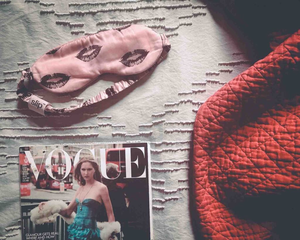 Flatlay of pink silk sleep mask, Vogue magazine, and red quilt — nighttime ritual aesthetic symbolizing rest, recovery, and quiet luxury.