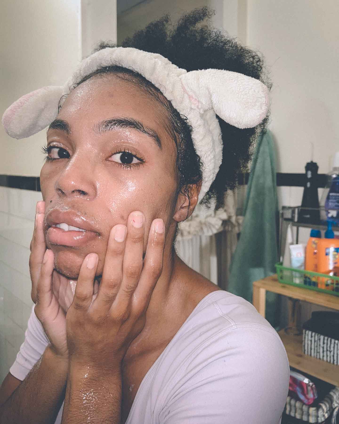 Woman with fluffy headband and dewy skin applying skincare in a cozy bathroom — soft self-care and morning ritual photography.