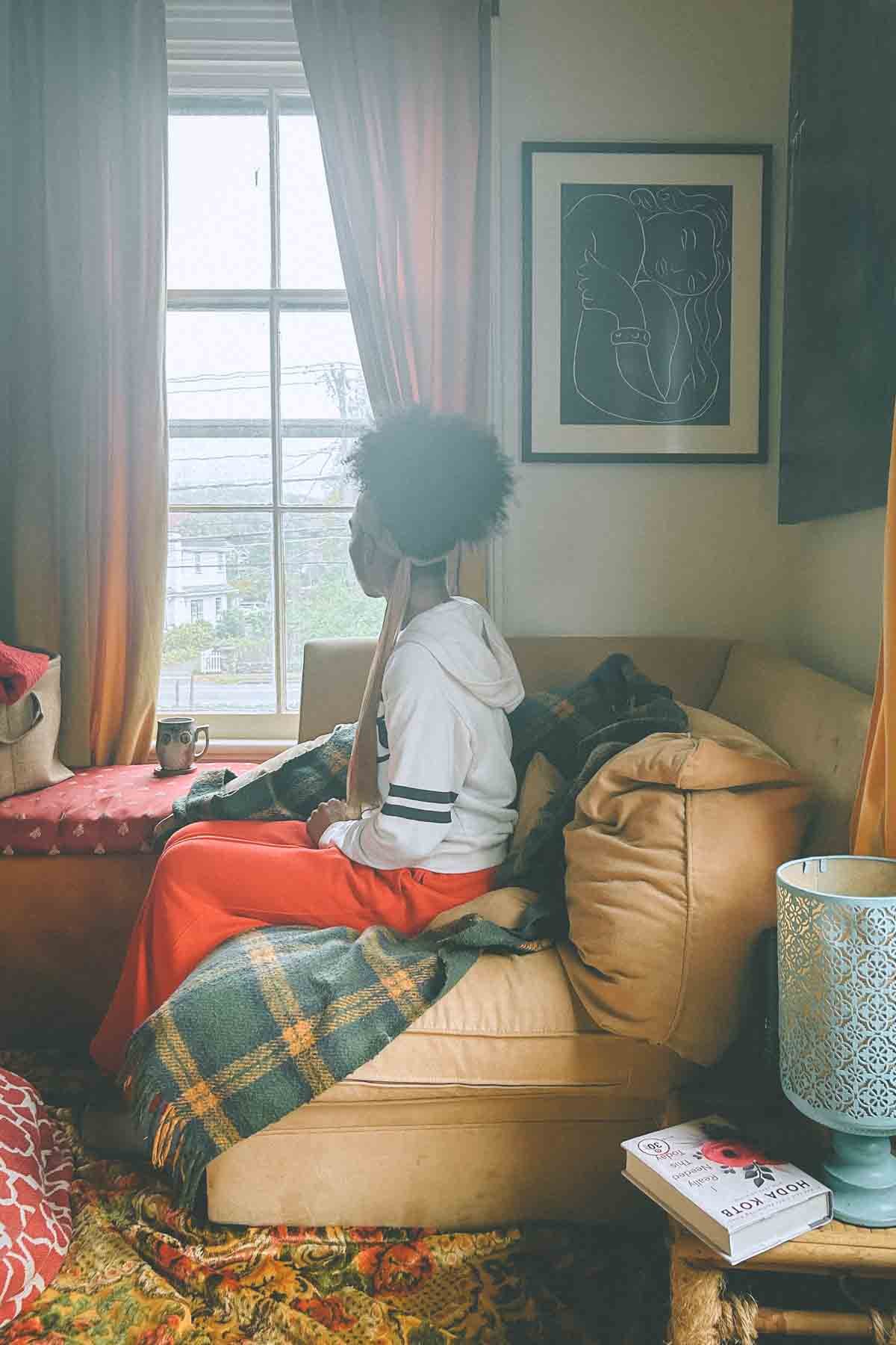 Woman gazing out a rain-streaked window in her apartment, symbolizing reflection and the quiet act of starting again.