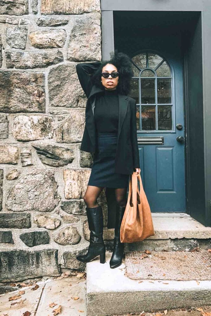 Bohemian fashion blogger wearing a black blazer, turtleneck, mini skirt, and tall leather boots while posing outside a stone building for a fall “Inside My Closet” outfit editorial.