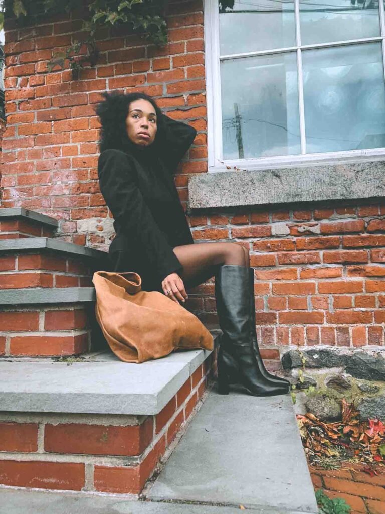 Boho-luxe blogger in a black mini skirt, blazer, and knee-high leather boots sitting on brick steps with a suede tote, fall outfit inspiration for Inside My Closet blog.
