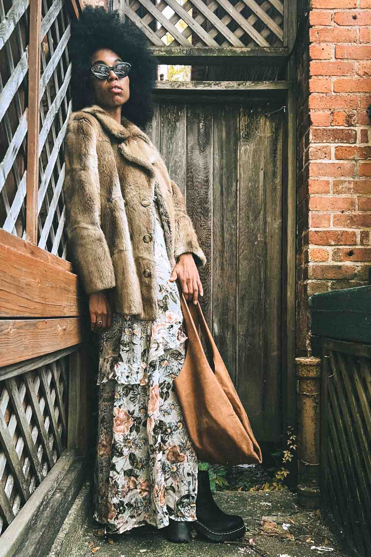 Bohemian woman wearing vintage fur coat over floral maxi dress, posing in a rustic courtyard for fall Inside My Closet feature.