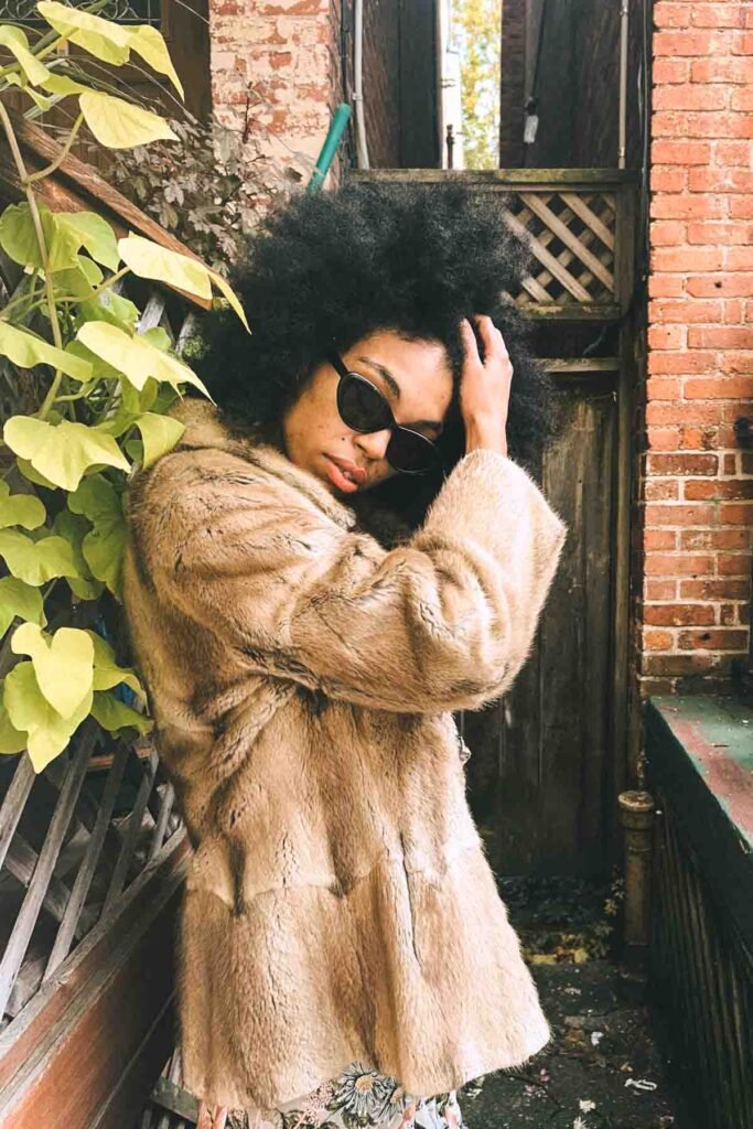 Editorial portrait of a bohemian blogger in a vintage faux-fur coat and cat-eye sunglasses, posing beside brick walls and leafy green vines.
