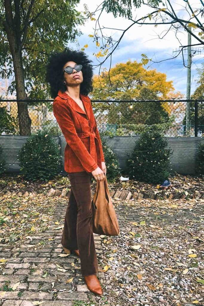 Bohemian blogger wearing a rust suede jacket and brown flared pants, standing on a cobblestone path surrounded by autumn leaves and trees in New York.