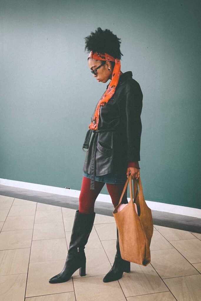 Bohemian fashion blogger wearing a black leather jacket, denim mini skirt, red tights, and an orange floral scarf, holding a suede tote bag against a muted green wall.