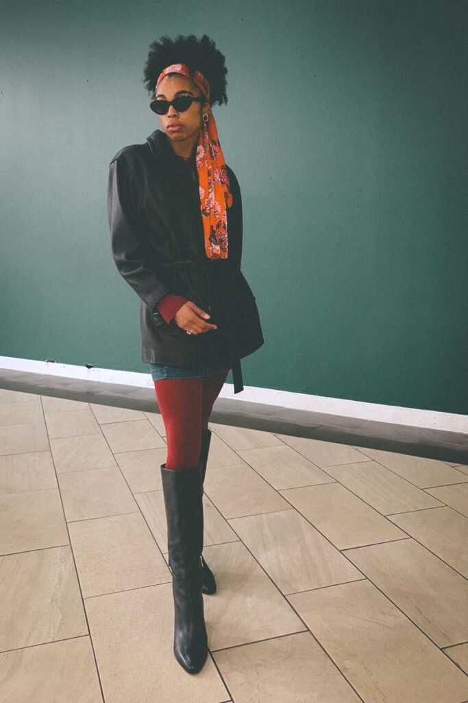 Bohemian fashion blogger wearing a belted black leather jacket, red tights, knee-high black boots, and an orange floral scarf posing against a teal wall.