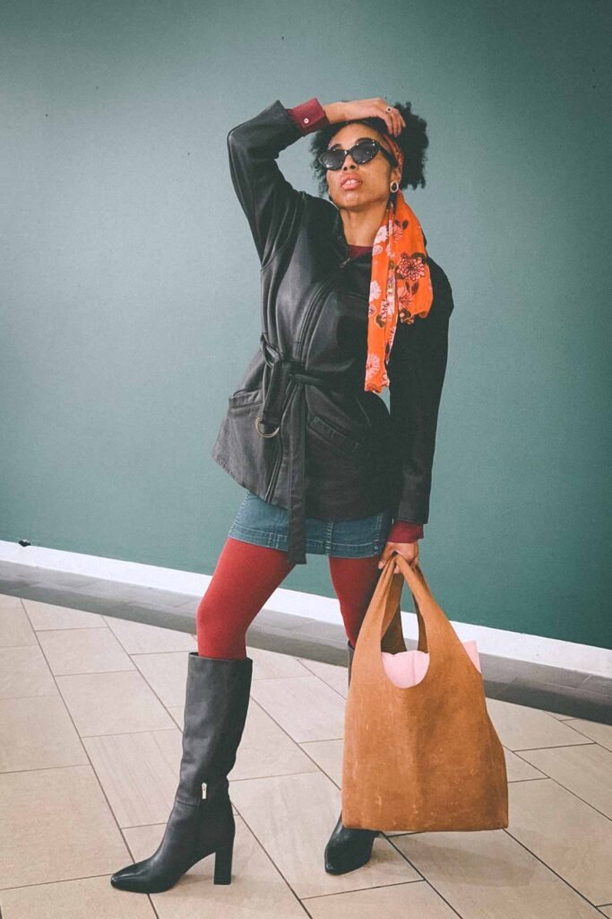 Bohemian blogger posing in a vintage-inspired fall outfit with a belted black leather jacket, denim skirt, red tights, and orange scarf, holding a tan suede tote.