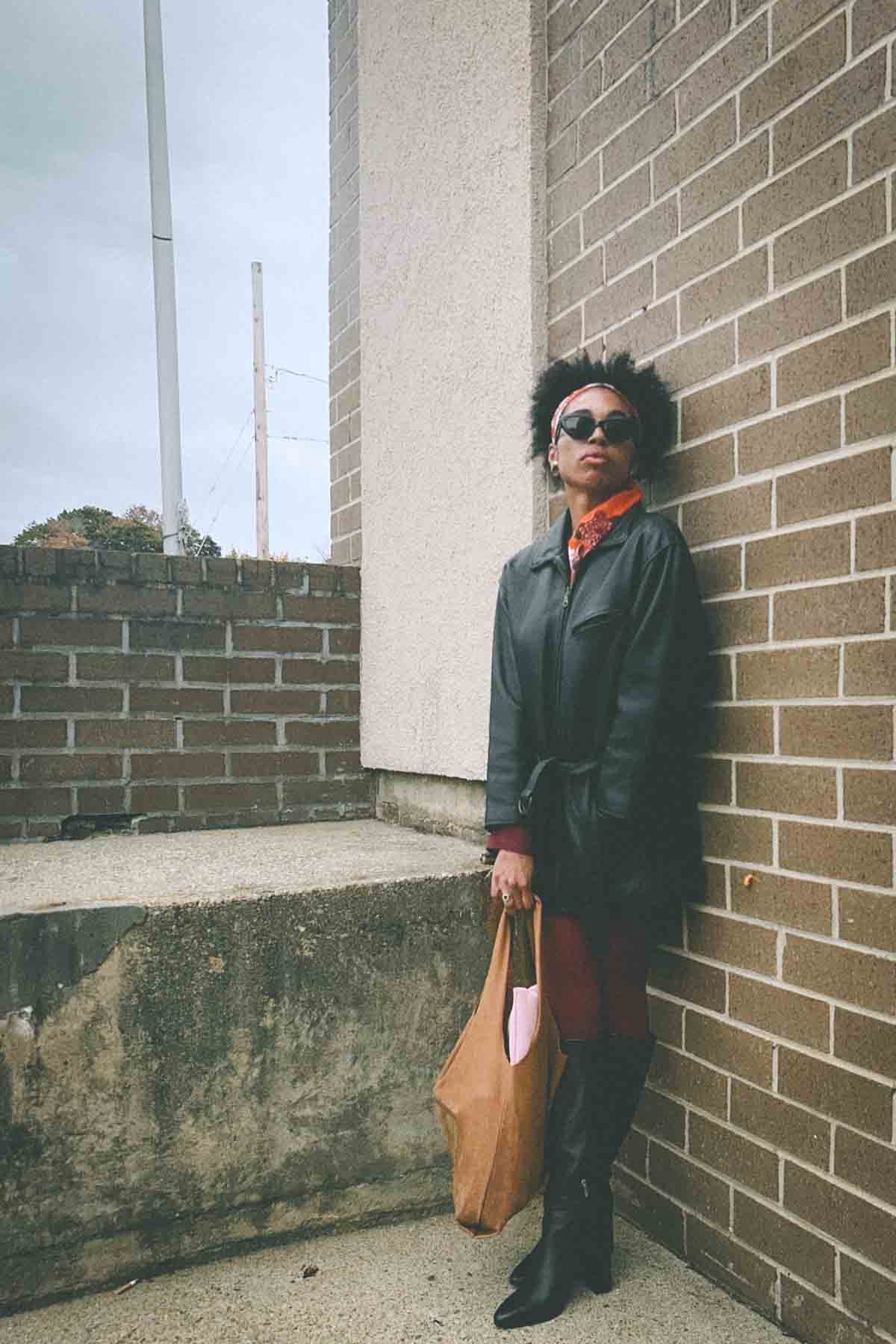 Bohemian fashion blogger leaning against brick wall in a belted black leather jacket, red tights, and tall boots, carrying a suede tote — fall street style inspiration.