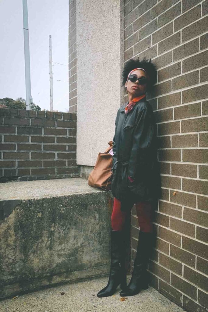 Bohemian fashion blogger wearing a belted black leather jacket, red tights, and tall black boots leaning against a brick wall with a brown suede tote.