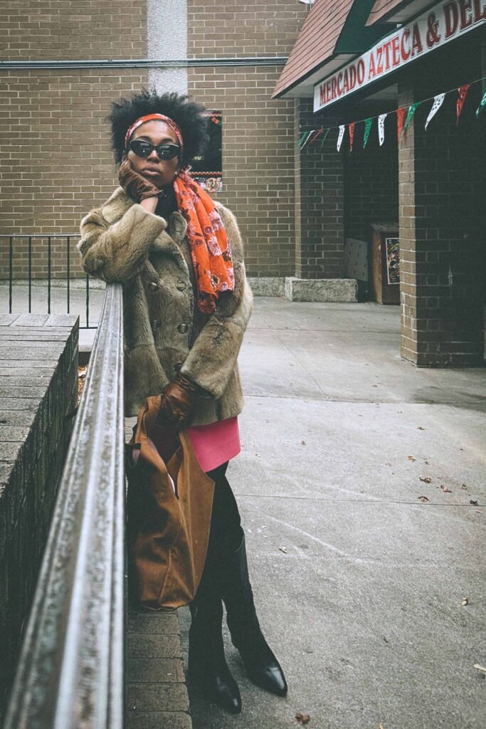 Bohemian fashion blogger in a vintage fur coat and orange floral scarf posing outside a storefront with tall black boots and a brown suede tote.