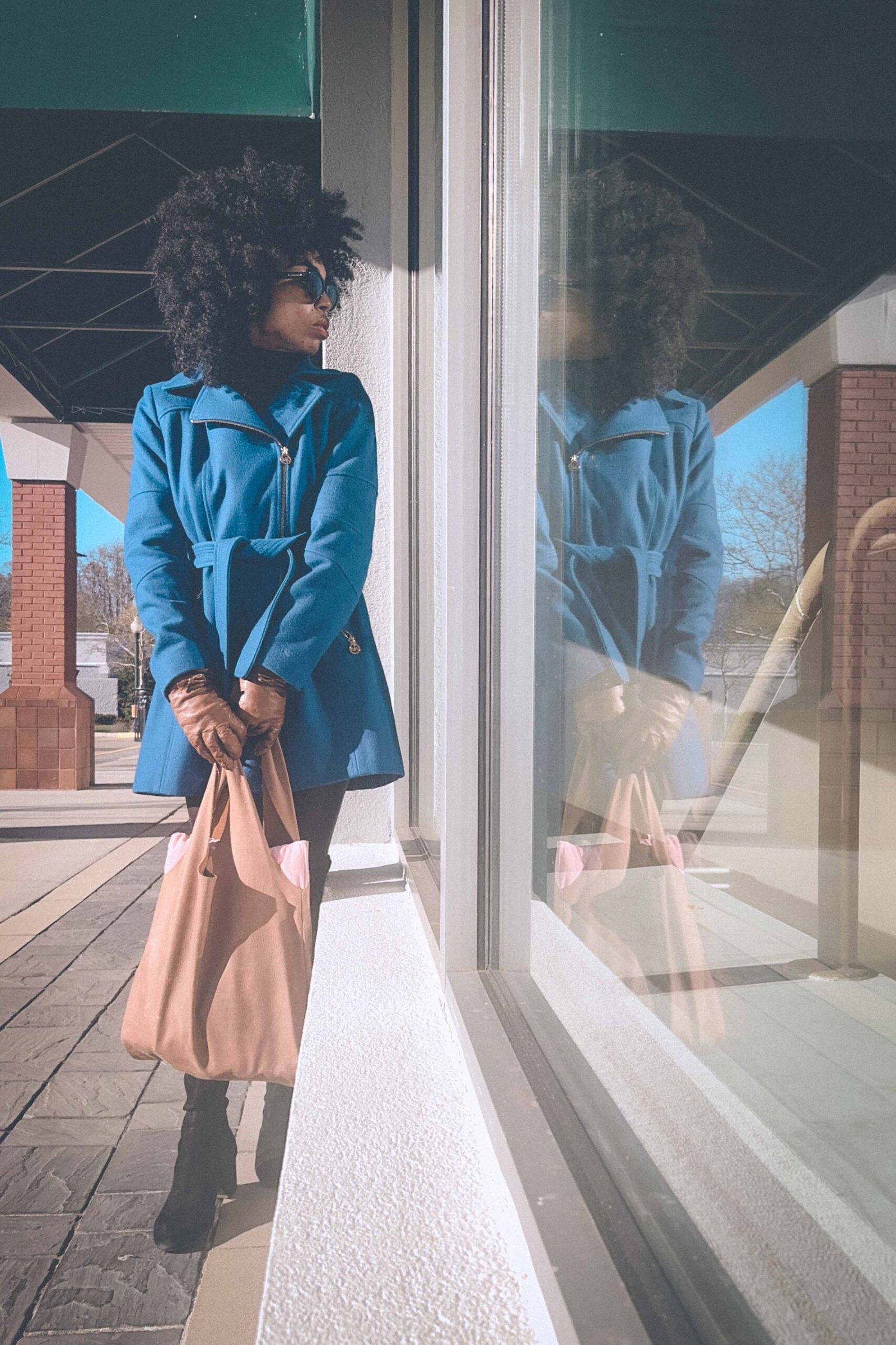 Boho fashion blogger in a blue winter coat standing by a reflective storefront window on a chilly day, embodying the mood of early winter blues.