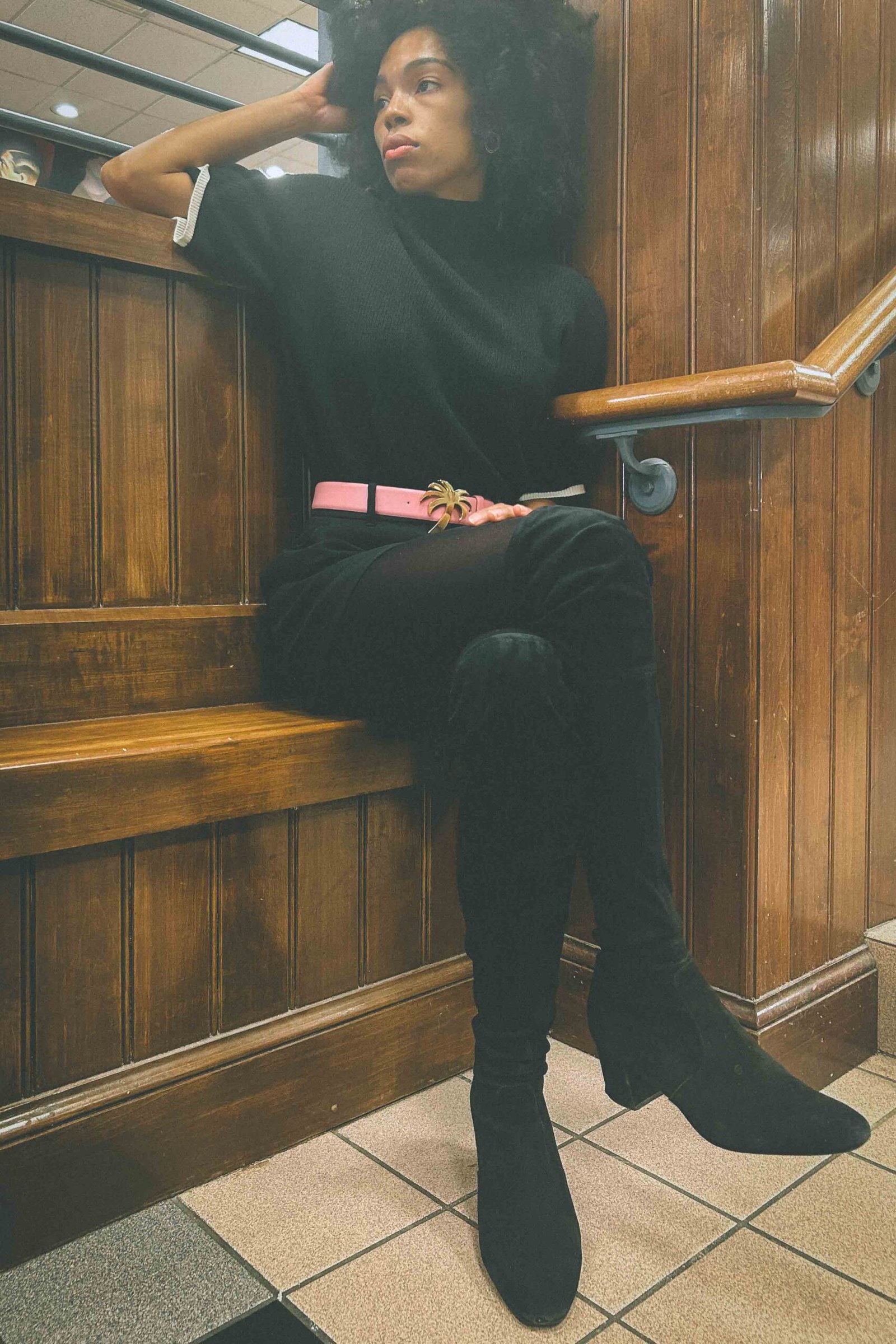 Moody editorial portrait of a woman sitting on wooden stairs in a mall, wearing an all-black outfit with a pink belt, capturing holiday sadness and introspection.