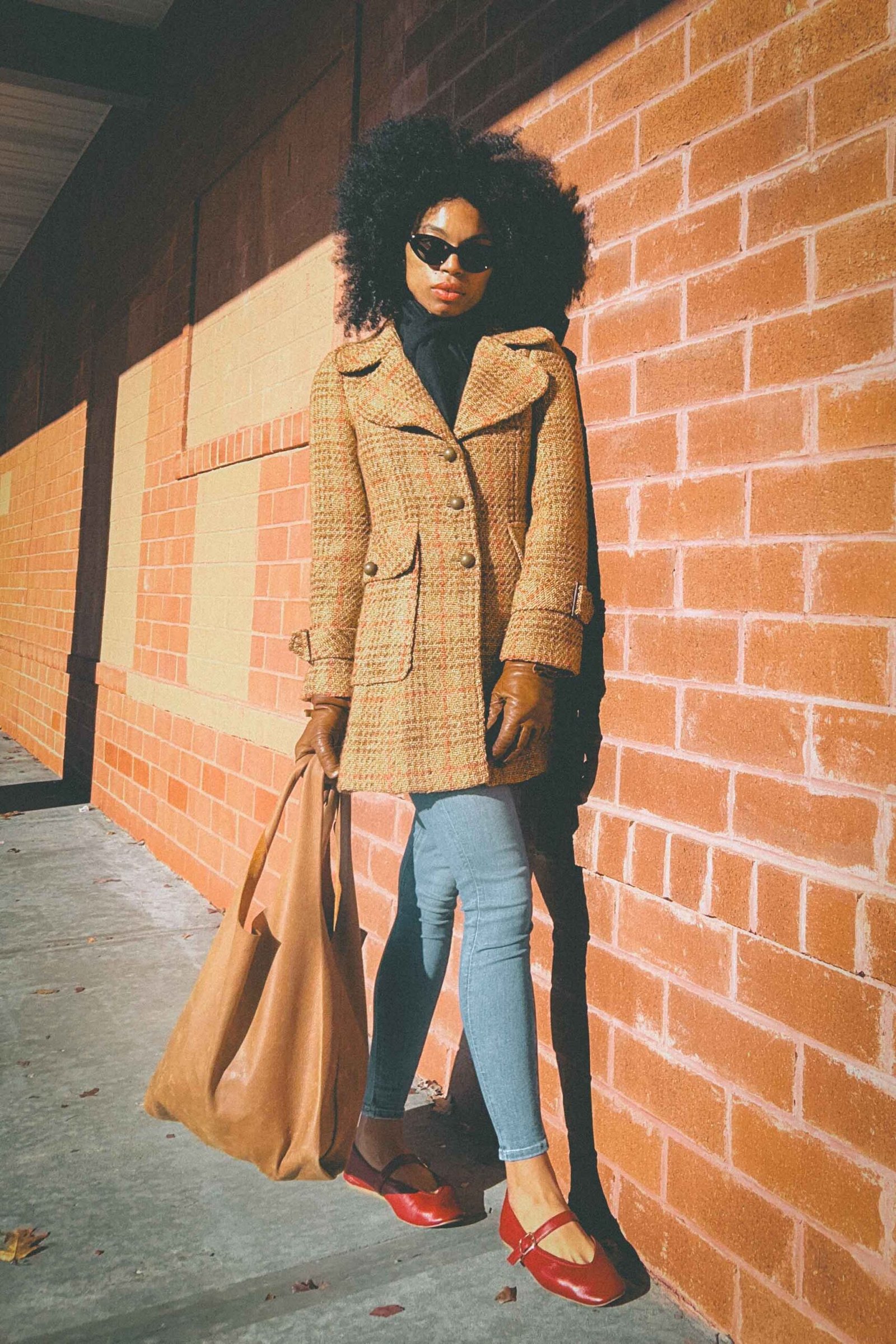 Boho fashion blogger standing against a sunlit brick wall wearing a vintage tweed coat, red Mary Jane flats, and oversized sunglasses during the early holiday season.